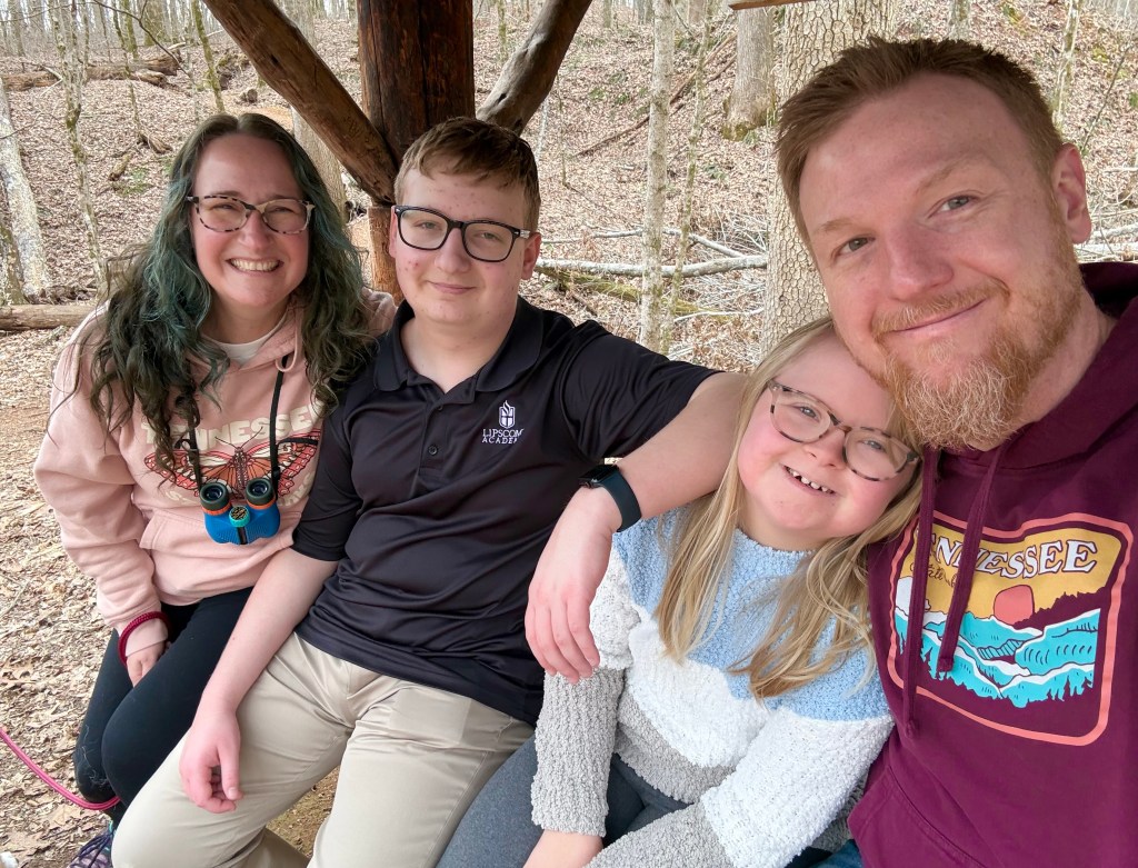 A light-skinned family of four is seated on an outdoor bench in winter: a woman with glasses and long, wavy dark hair with green highlights, a teenage boy wearing a dark polo and glasses, a blonde girl with glasses who has Down syndrome, and a man with red hair and beard.